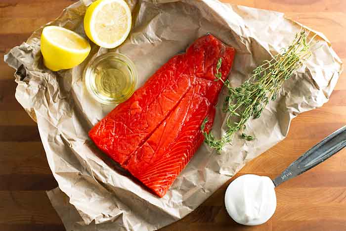 Raw salmon fillet, lemon halves, fresh thyme, olive oil in a small bowl, and sour cream on brown paper