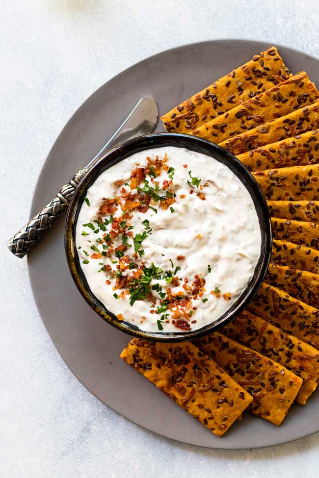 Onion dip garnished with bacon and parsley on a plate with crackers