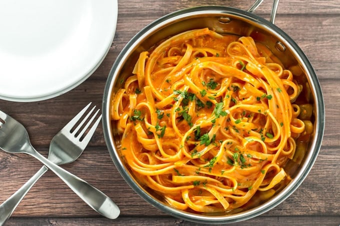 A skillet with roasted red pepper alfredo with forks and plates on the side