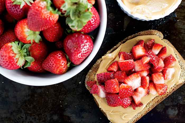 a slice of toast topped with peanut butter, diced strawberries, with a bowl of whole strawberries next to it