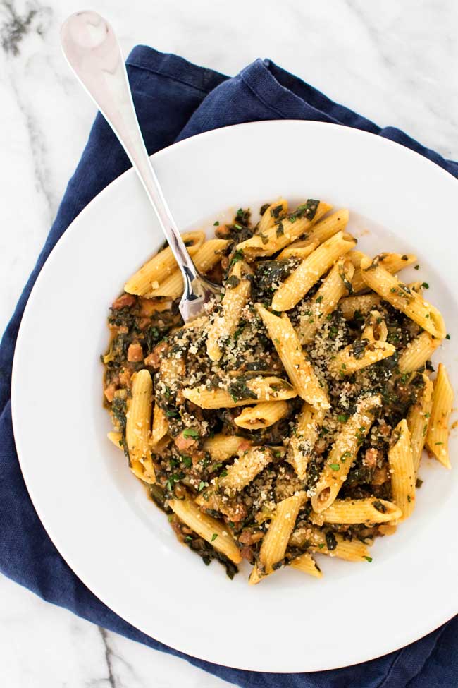 spinach penne with pancetta in a white bowl with a fork and a blue napkin