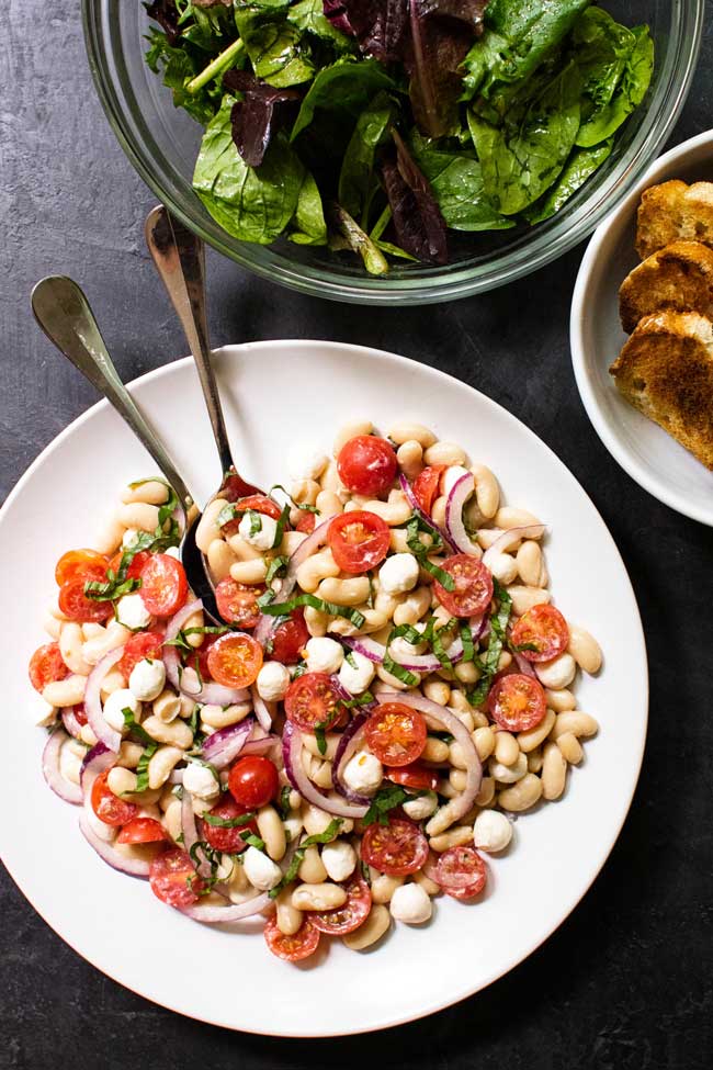 White Bean Tomato Salad on a white plate with a bowl of lettuce and bread on the side