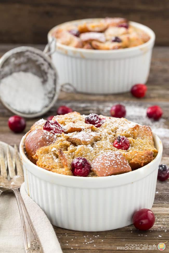 Two Overnight Eggnog Cranberry French Toast servings in individual white ramekins dusted with powdered sugar with some cranberries scattered around and powdered sugar in a sieve in the background.