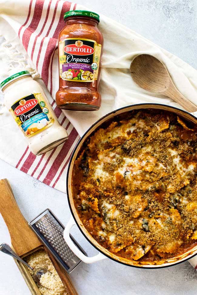 Pan with baked ravioli in spinach rosa sauce with bottles of bertolli sauces, grated parmesan cheese, and a wooden spoon