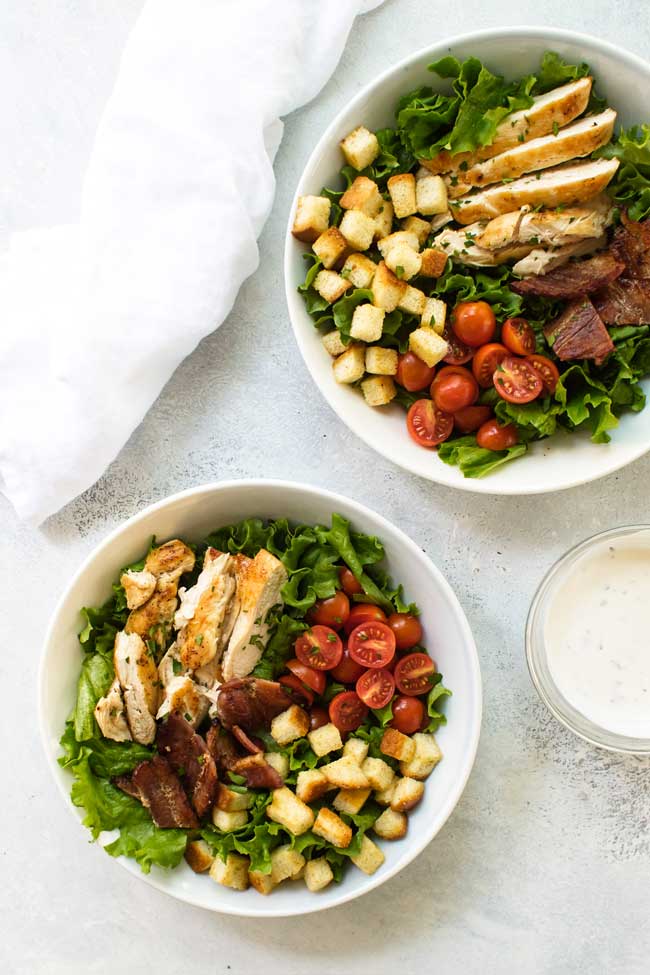 Two bowls with lettuce, cooked chicken, tomatoes, bacon, homemade croutons and lettuce with dressing in a small bowl on the side