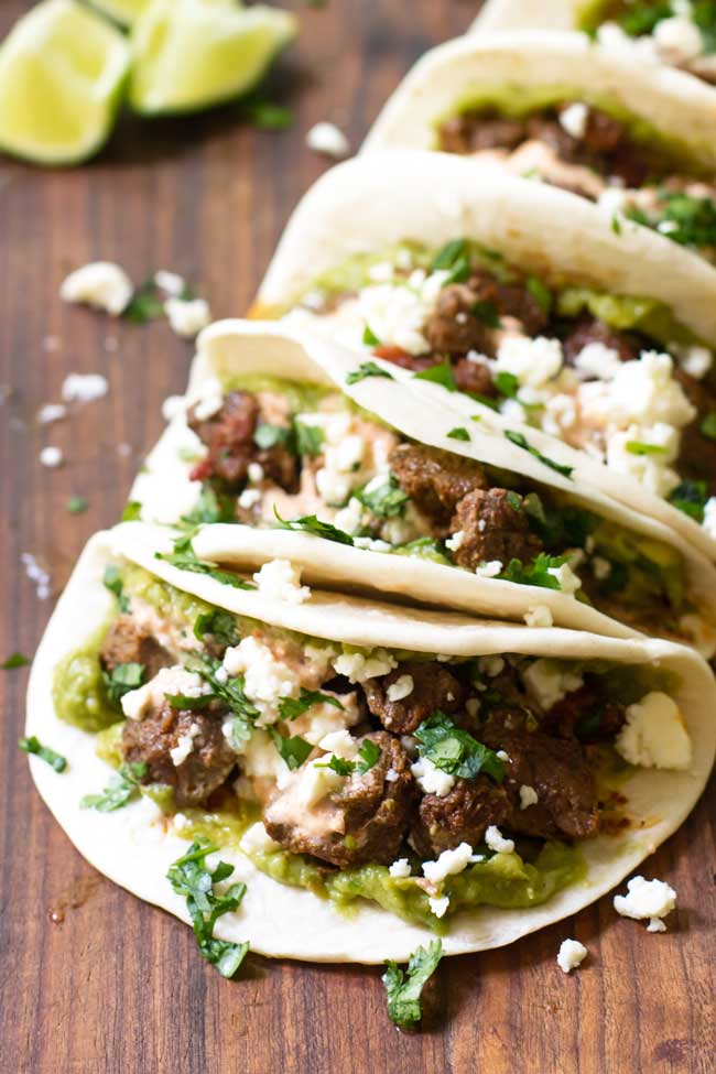 four braised beef tacos lined up on a cutting board and garnished with feta cheese, cilantro, and a creamy salsa sauce