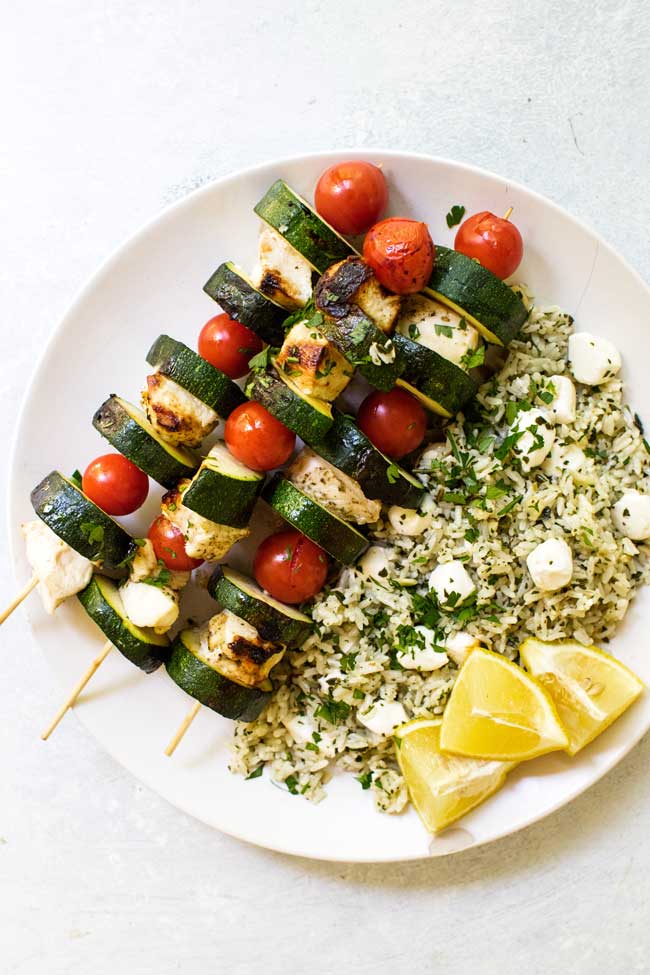 chicken kabobs on a white plate with pesto rice and lemon wedges