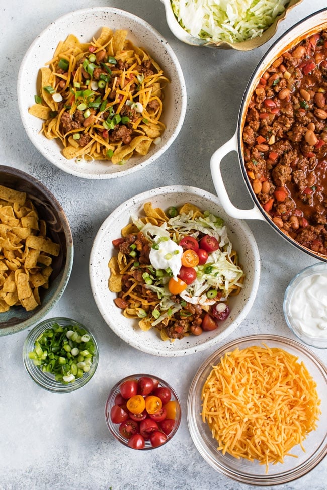 Overhead photo of a frito pies in bowls surrounded by frito pie toppings