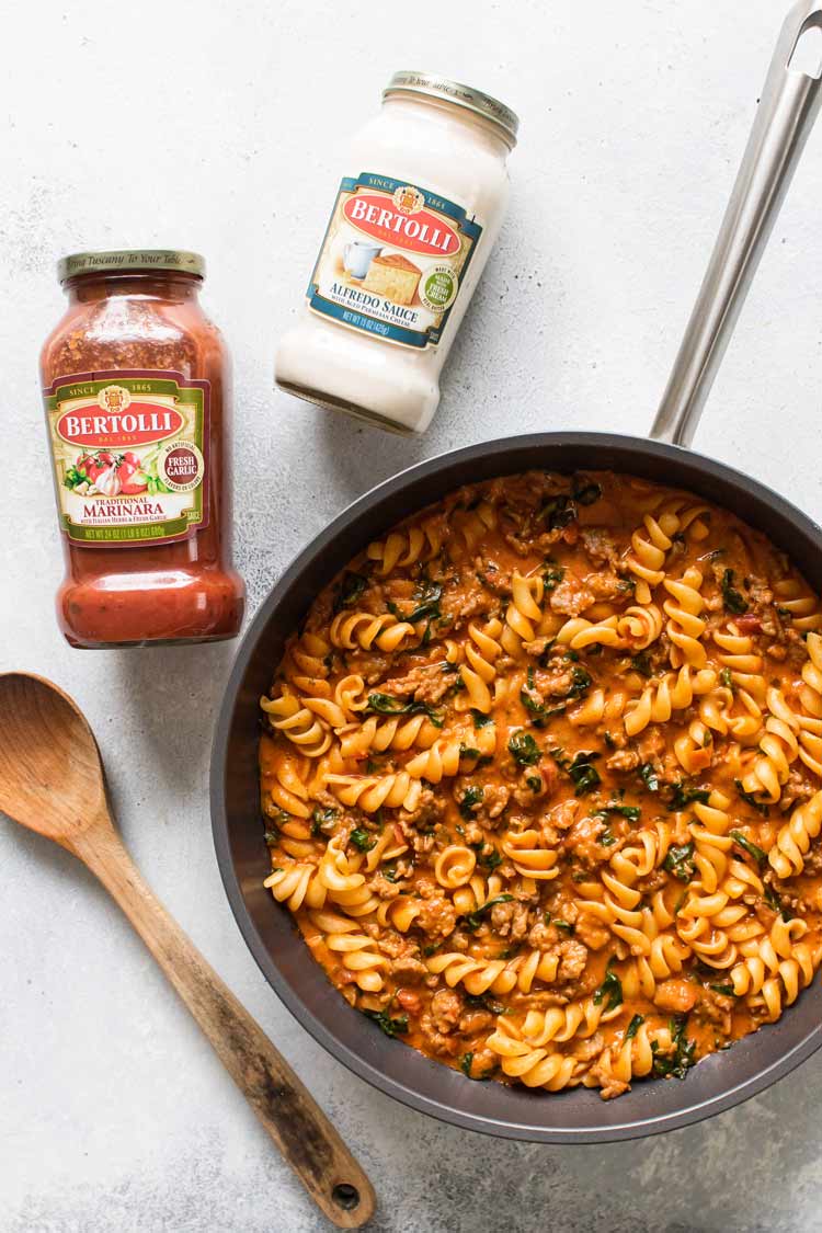 overhead photo of a skillet of pasta with two jars of sauce and a wooden spoon