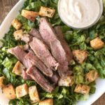 a platter with lettuce and topped with croutons, sliced steak, and a small bowl of caesar dressing.