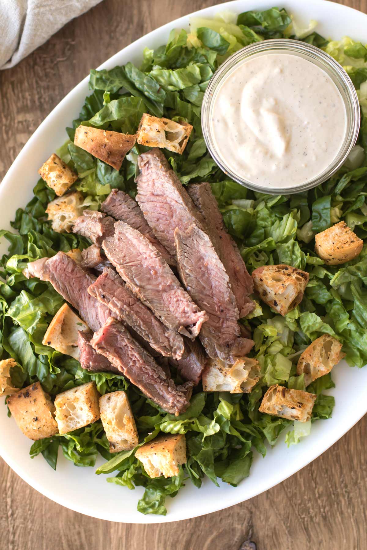 a platter with lettuce and topped with croutons, sliced steak, and a small bowl of caesar dressing.