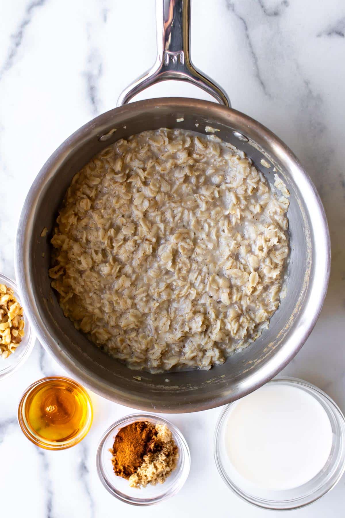 the cooked oatmeal in a pan with the milk, sugar, cinnamon, honey, and walnuts in bowls around it.