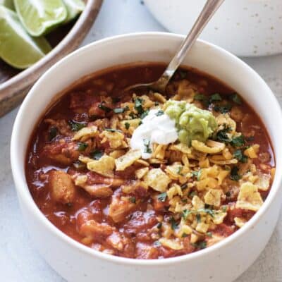 a bowl of red chicken chili topped with fritos, sour cream, and guacamole.
