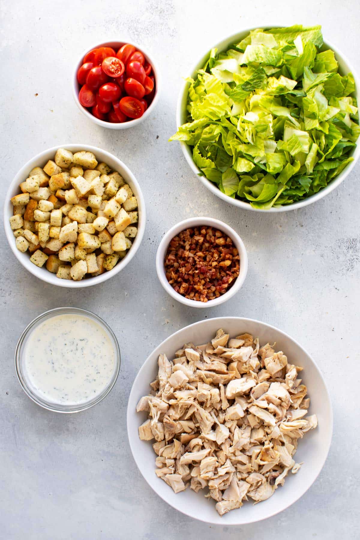 bowls of lettuce, tomatoes, croutons, bacon bits, dressing, and chopped cooked chicken.